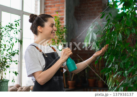 Running of own business. Young woman florist with spray bottle watering fertilizing plants in botanical store. Small business owner working at flower shop smiling surrounded by plants Small business 137938137