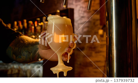 Draft beer overflowing glass with thick foam in bartender hand dark moody bar. 137939732