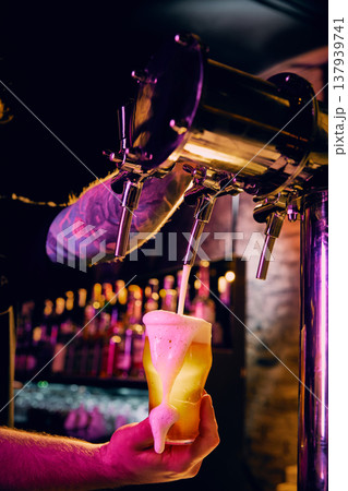 Draft beer pouring into glass with colorful neon lighting and foam overflow in bar. 137939741
