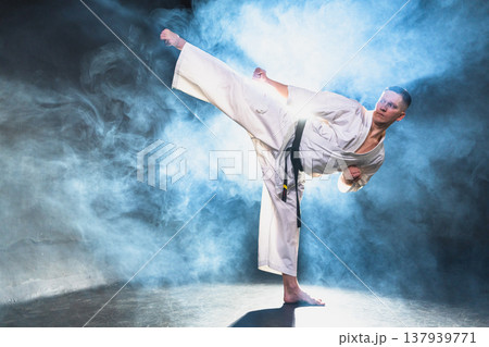 Strong man fighter in white kimono for karate martial arts with black belt preparing for fight training in black background. Man training moves punches kicks practicing shadow fight ready for sparring Strong man fighter in white kimono for karate martial arts with black belt preparing for fight training in black background. Man training moves punches kicks practicing shadow fight ready for sparring 137939771