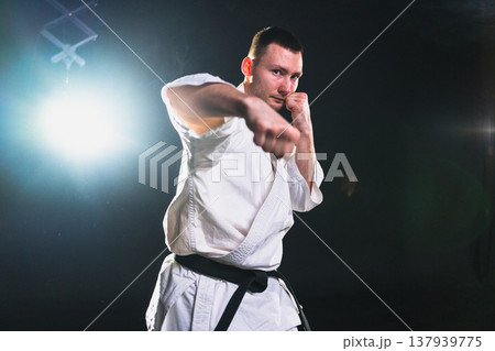 Strong man fighter in white kimono for karate martial arts with black belt preparing for fight training in black background. Man training moves punches kicks practicing shadow fight ready for sparring Strong man fighter in white kimono for karate martial arts with black belt preparing for fight training in black background. Man training moves punches kicks practicing shadow fight ready for sparring 137939775