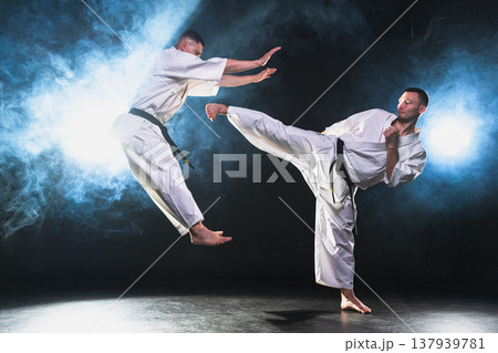 Martial arts masters on dark background. Strong man fighters in white kimono for karate martial arts fighting training in black gym. Karate fighters training punches kicks practicing fight sparring Martial arts masters on dark background. Strong man fighters in white kimono for karate martial arts fighting training in black gym. Karate fighters training punches kicks practicing fight sparring 137939781