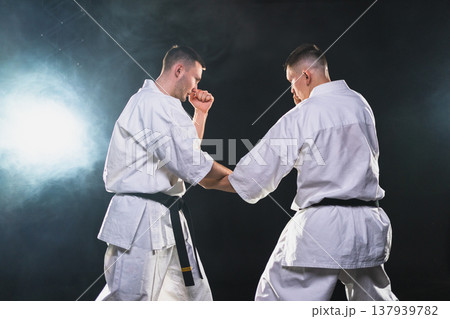 Martial arts masters on dark background. Strong man fighters in white kimono for karate martial arts fighting training in black gym. Karate fighters training punches kicks practicing fight sparring Martial arts masters on dark background. Strong man fighters in white kimono for karate martial arts fighting training in black gym. Karate fighters training punches kicks practicing fight sparring 137939782