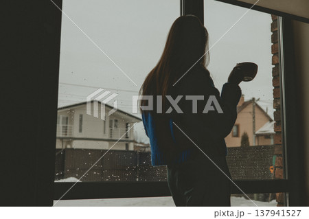 Pensive silhouette of a woman holding a coffee mug, looking out a large window at a snowy suburban winter landscape. Rainy gloomy weather. Pensive silhouette of a woman holding a coffee mug, looking out a large window at a snowy suburban winter landscape. Rainy gloomy weather. 137941527