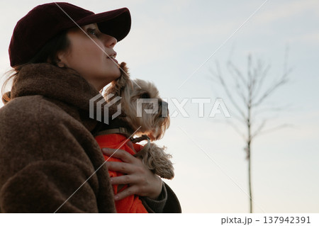 Candid portrait of a woman in a maroon cap and brown fleece jacket holding a small dog in an orange sweater. Lifestyle pet photography, emotional bond Candid portrait of a woman in a maroon cap and brown fleece jacket holding a small dog in an orange sweater. Lifestyle pet photography, emotional bond 137942391