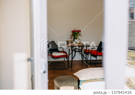 Cozy studio apartment interior with a dining table, red chairs, and fresh tulips. Modern minimalist decor, natural light, lifestyle home atmosphere Cozy studio apartment interior with a dining table, red chairs, and fresh tulips. Modern minimalist decor, natural light, lifestyle home atmosphere 137942416