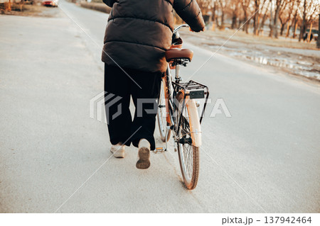 Back view of a woman walking with a vintage brown bicycle on a city or suburb road. Autumn or spring season, urban transportation. Lifestyle scene. Back view of a woman walking with a vintage brown bicycle on a city or suburb road. Autumn or spring season, urban transportation. Lifestyle scene. 137942464