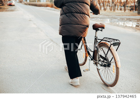 Back view of a woman walking with a vintage brown bicycle on a city or suburb road. Autumn or spring season, urban transportation. Lifestyle scene. 137942465