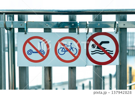 Red prohibition signs on a wooden fence near a waterfront, forbidding electric scooters, bicycles, swimming. Urban safety regulations and public rules in a recreational area or pedestrian zone. Red prohibition signs on a wooden fence near a waterfront, forbidding electric scooters, bicycles, swimming. Urban safety regulations and public rules in a recreational area or pedestrian zone. 137942828