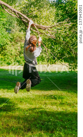Young woman hanging from a tree branch in a summer park. Active girl playing outdoors in nature. Fun leisure activity, childhood joy, and summer adventure concept for recreation. 137943001