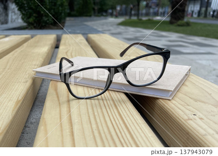 Glasses rest on a light-colored notebook on a wooden table in a park during the day Glasses rest on a light-colored notebook on a wooden table in a park during the day 137943079