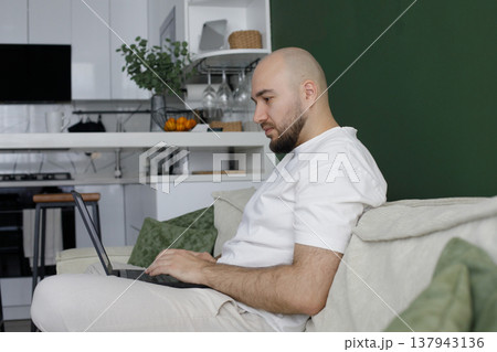 Handsome man working on laptop at home while sitting on sofa Handsome man working on laptop at home while sitting on sofa 137943136