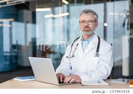 Senior male doctor with eyeglasses and stethoscope sitting at his desk, typing on a laptop, providing professional medical consultation and healthcare services in a clinic environment 137943416