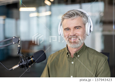Smiling mature man broadcasting audio content with a professional studio microphone and headphones, showcasing modern communication, streaming, and entertainment concepts Smiling mature man broadcasting audio content with a professional studio microphone and headphones, showcasing modern communication, streaming, and entertainment concepts 137943423