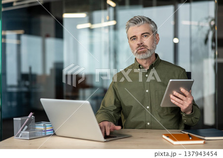 Mature businessman with gray hair and beard sits at a modern office desk, using laptop and tablet for remote work and online communication, focused on planning and strategy 137943454