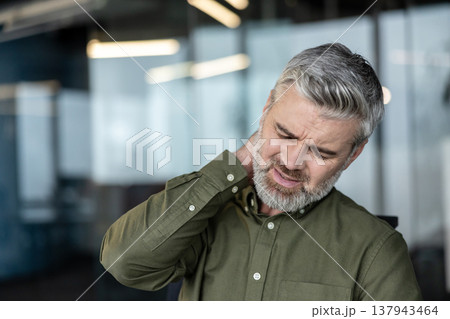 Mature businessman with gray hair and beard rubbing his neck in a modern office, grimacing from cervical pain and poor posture while working at a computer, stressed and fatigued 137943464