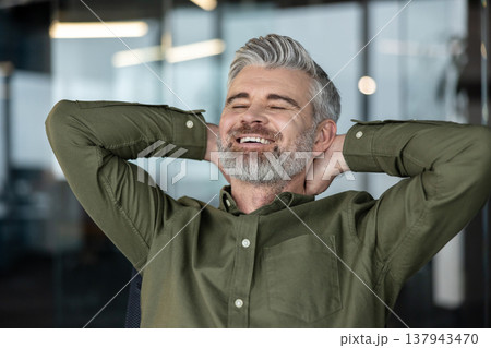Mature man with grey hair and beard leaning back with hands behind his head, smiling with eyes closed, embodying business success, relaxation, and satisfaction in a modern office environment Mature man with grey hair and beard leaning back with hands behind his head, smiling with eyes closed, embodying business success, relaxation, and satisfaction in a modern office environment 137943470