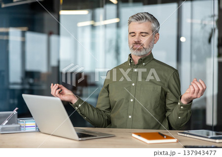 Mature man finding zen, meditating with closed eyes and mudra gesture at his desk with a laptop, practicing mindfulness to relieve stress and boost focus in a modern office environment Mature man finding zen, meditating with closed eyes and mudra gesture at his desk with a laptop, practicing mindfulness to relieve stress and boost focus in a modern office environment 137943477