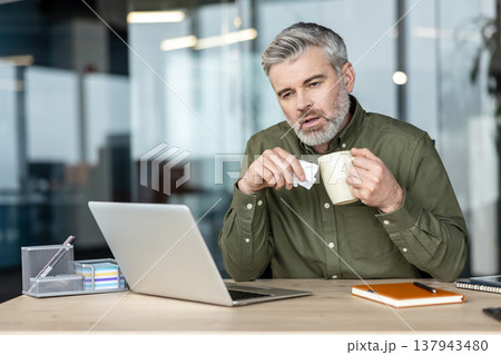 Mature man sitting at his office desk, holding a tissue and a mug, experiencing symptoms of a cold or flu while trying to continue working, highlighting workplace presenteeism Mature man sitting at his office desk, holding a tissue and a mug, experiencing symptoms of a cold or flu while trying to continue working, highlighting workplace presenteeism 137943480