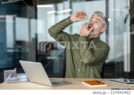 Mature businessman applying moisturizing eye drops to alleviate dry eyes and computer vision syndrome caused by prolonged screen time while working in modern office environment 137943482