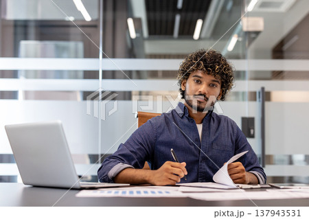 Portrait of a serious young Indian businessman working in the office with a laptop and documents, looking at the camera. 137943531