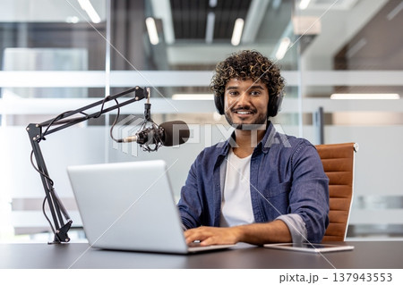 Portrait of a young Indian man sitting in an office at a desk with a microphone stand and looking at the camera with a smile. Portrait of a young Indian man sitting in an office at a desk with a microphone stand and looking at the camera with a smile. 137943553