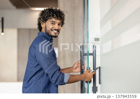 Portrait of a young Indian man standing in an office near the front door and opening it with a phone app. 137943559
