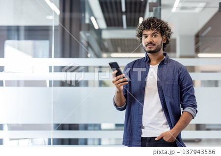 Portrait of a young Indian man standing in an office, holding his hand in his pocket, using his phone and looking at the camera. Portrait of a young Indian man standing in an office, holding his hand in his pocket, using his phone and looking at the camera. 137943586