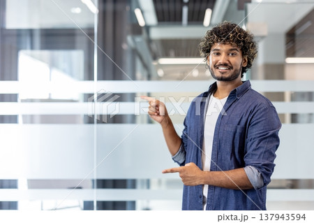 Portrait of a smiling Indian young man standing in the office and pointing with his fingers at an empty space. 137943594