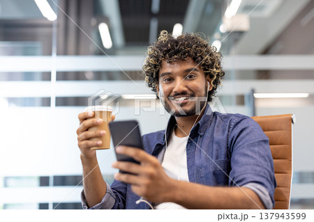 Close-up portrait of a young Indian man wearing headphones, holding a phone and a cup of coffee in his hands, smiling and looking at the camera. 137943599