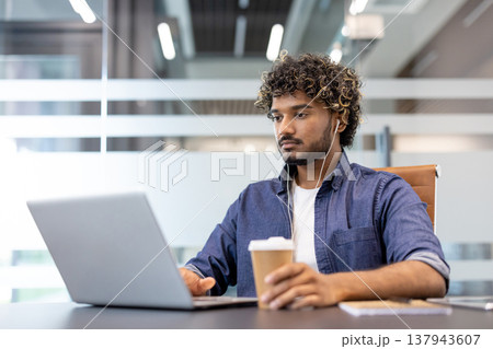 Serious and focused young Indian man working with headphones on a laptop in the office, sitting in a chair, drinking coffee. Serious and focused young Indian man working with headphones on a laptop in the office, sitting in a chair, drinking coffee. 137943607