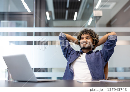 A young Indian man is satisfied with the final result, sitting in an office chair, hands behind his head, and resting. A young Indian man is satisfied with the final result, sitting in an office chair, hands behind his head, and resting. 137943748