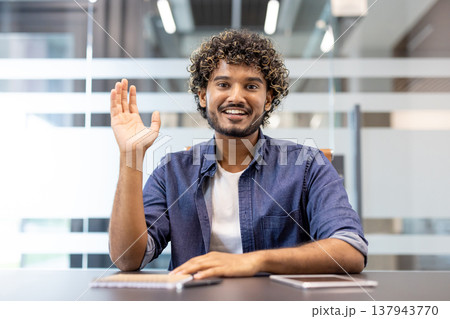 Close-up portrait of a young smiling Indian man sitting in an office at a desk facing the camera and raising his right hand. Close-up portrait of a young smiling Indian man sitting in an office at a desk facing the camera and raising his right hand. 137943770