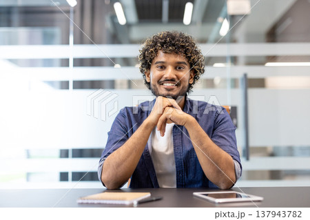 Portrait of a smiling young Indian man sitting at a desk in an office facing the camera. Portrait of a smiling young Indian man sitting at a desk in an office facing the camera. 137943782