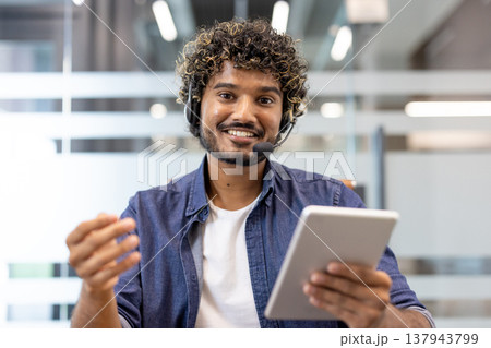 Close-up photo of a young Indian man in a headset looking and mumbling at the camera, taking notes in a notebook. 137943799
