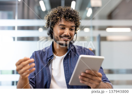 Muslim young man talking to camera in office, wearing headset and using tablet. 137943800