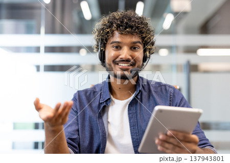 Portrait of a young smiling Indian man in a headset sitting at a desk in the office in front of the camera and talking on a video call. Portrait of a young smiling Indian man in a headset sitting at a desk in the office in front of the camera and talking on a video call. 137943801