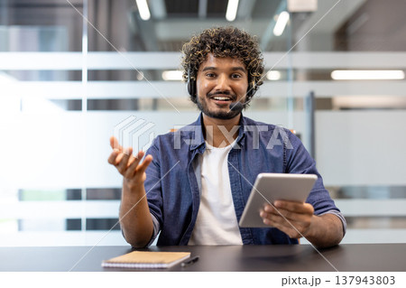 Portrait of a smiling Indian man in a headset sitting in the office in front of the camera and talking, holding a tablet in his hand. Portrait of a smiling Indian man in a headset sitting in the office in front of the camera and talking, holding a tablet in his hand. 137943803