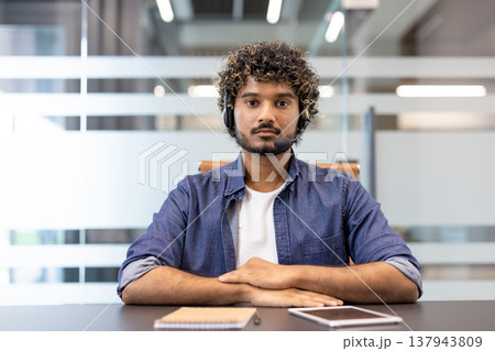 Photo of a young Indian man wearing headphones sitting at a desk in an office facing the camera and making a video call. Photo of a young Indian man wearing headphones sitting at a desk in an office facing the camera and making a video call. 137943809