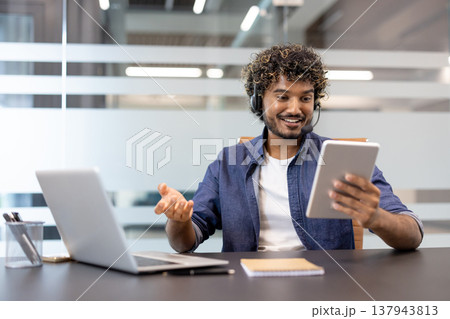 A young Indian man in a suit works in an office at a laptop and has an online conversation via a tablet he holds in his hand. A young Indian man in a suit works in an office at a laptop and has an online conversation via a tablet he holds in his hand. 137943813