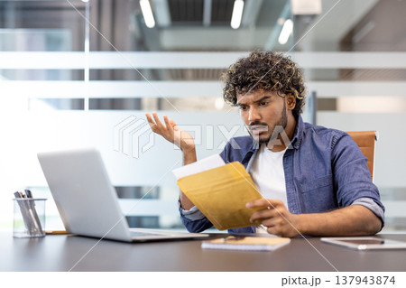 A worried young Indian man sits at his desk in the office and looks disappointedly at the letter he received. A worried young Indian man sits at his desk in the office and looks disappointedly at the letter he received. 137943874
