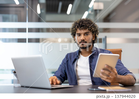 Portrait of a young serious Indian man sitting in the office at a desk with a laptop. holding a tablet in his hand and looking at the camera. 137944020