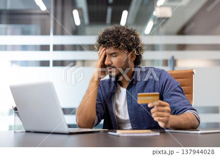 Shocked young Indian man sitting at a desk in the office, holding his head in his hands and holding a credit card. Shocked young Indian man sitting at a desk in the office, holding his head in his hands and holding a credit card. 137944028