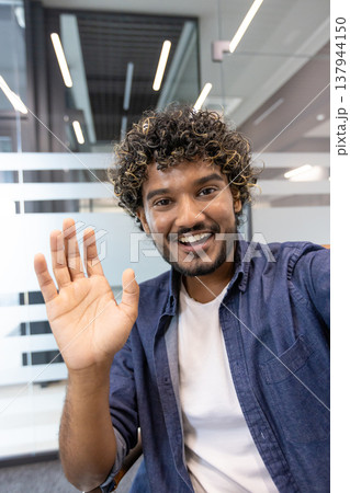 Vertical close-up photo of a young Indian man in an office talking to a camera he is holding in his hand. Vertical close-up photo of a young Indian man in an office talking to a camera he is holding in his hand. 137944150