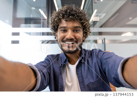 Close-up portrait of a young Indian man in an office, smiling and talking to the camera he is holding. Close-up portrait of a young Indian man in an office, smiling and talking to the camera he is holding. 137944153
