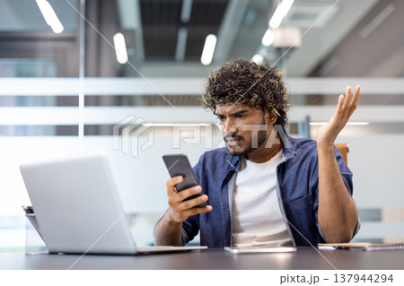 Worried young Muslim man looking at phone screen, sitting at desk in office and spreading hands in frustration. Worried young Muslim man looking at phone screen, sitting at desk in office and spreading hands in frustration. 137944294