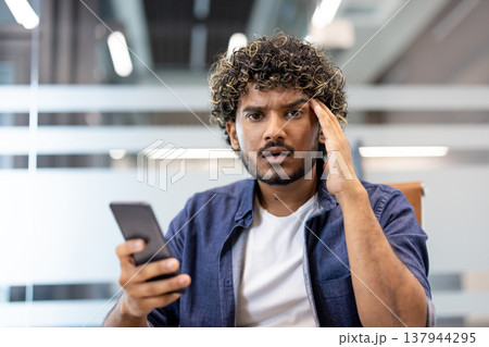 Close-up photo of a young Indian man holding a phone in his hand, standing in an office and looking worriedly into the camera. 137944295