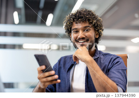Close-up portrait of a young Indian man sitting in a chair at a desk in an office, holding a phone and smiling at the camera. Close-up portrait of a young Indian man sitting in a chair at a desk in an office, holding a phone and smiling at the camera. 137944390