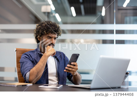 A serious and thoughtful young Indian man is sitting at his desk in the office and looking thoughtfully at his mobile phone screen. A serious and thoughtful young Indian man is sitting at his desk in the office and looking thoughtfully at his mobile phone screen. 137944408
