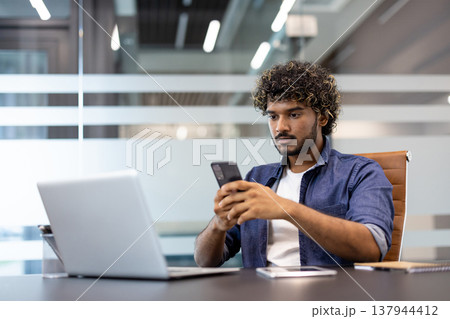Serious young Indian man sitting in the lobby at his desk and using his mobile phone. Serious young Indian man sitting in the lobby at his desk and using his mobile phone. 137944412